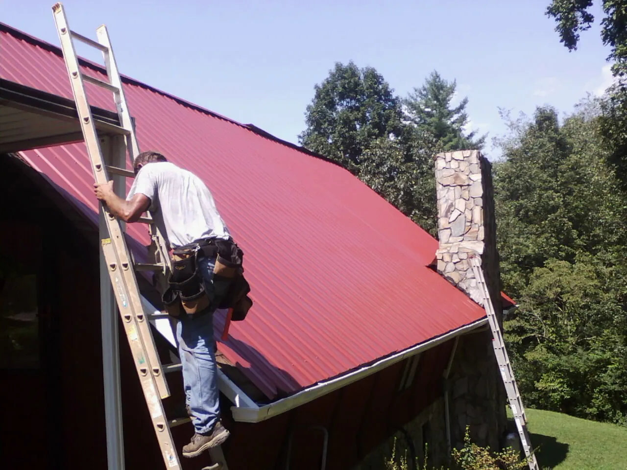 Roofer on a ladder beside a red metal roof and stone chimney — Soffit & Fascia Repair by Roofing Brothers in Steep Falls
