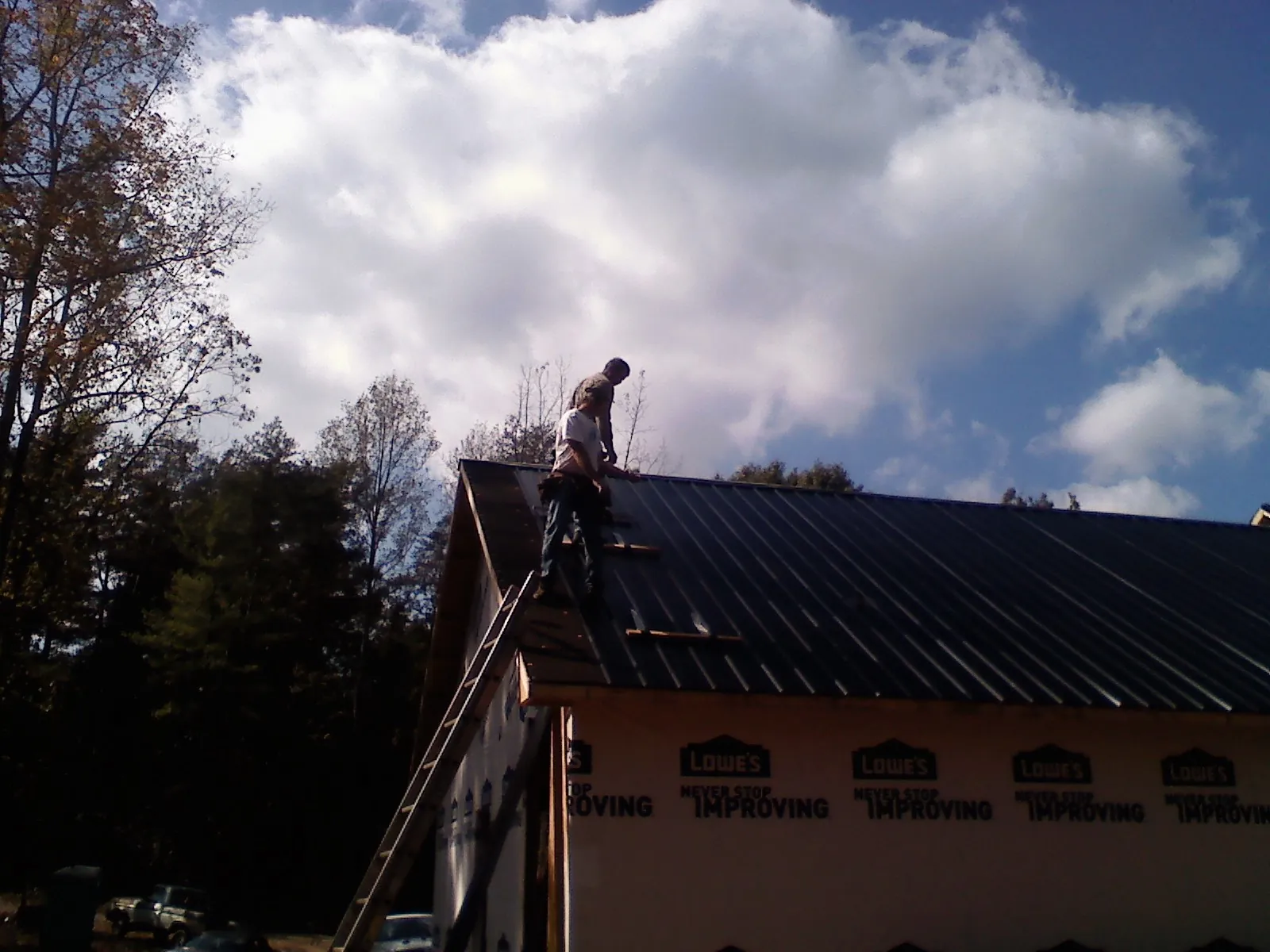Roofer fastening dark metal panels high against a cloudy sky, from a Roof Inspection job in Steep Falls